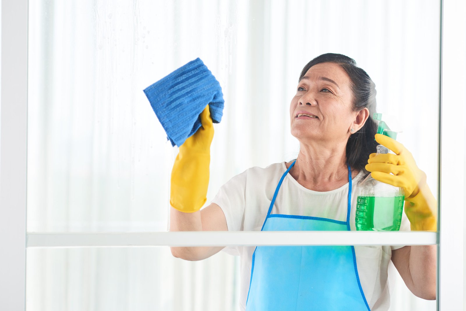 middle aged housekeeper wiping window with spray cleaner scaled